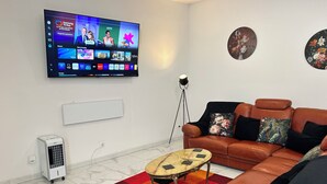 A living room with a flat-screen TV displaying a streaming service interface, a brown leather sofa with floral cushions, a small round coffee table, and a colorful checkered rug. - 1712-Villa Carre Sleeps4 end of terrace town house (Le Dorat)