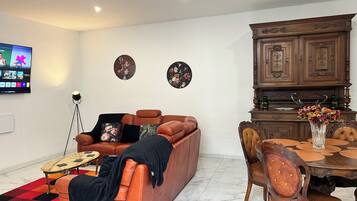 A living room with a brown leather sofa, a round wooden dining table with chairs, a TV mounted on the wall, and two wall-mounted decorative plates.