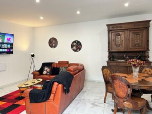 Ein Wohnzimmer mit braunem Ledersofa, einem runden Esstisch mit Stühlen, einem an der Wand befestigten Fernseher und zwei Wanddekorationen.