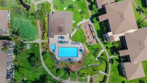Vista aérea de un complejo turístico con una piscina central, rodeado de céspedes cuidados y palmeras.