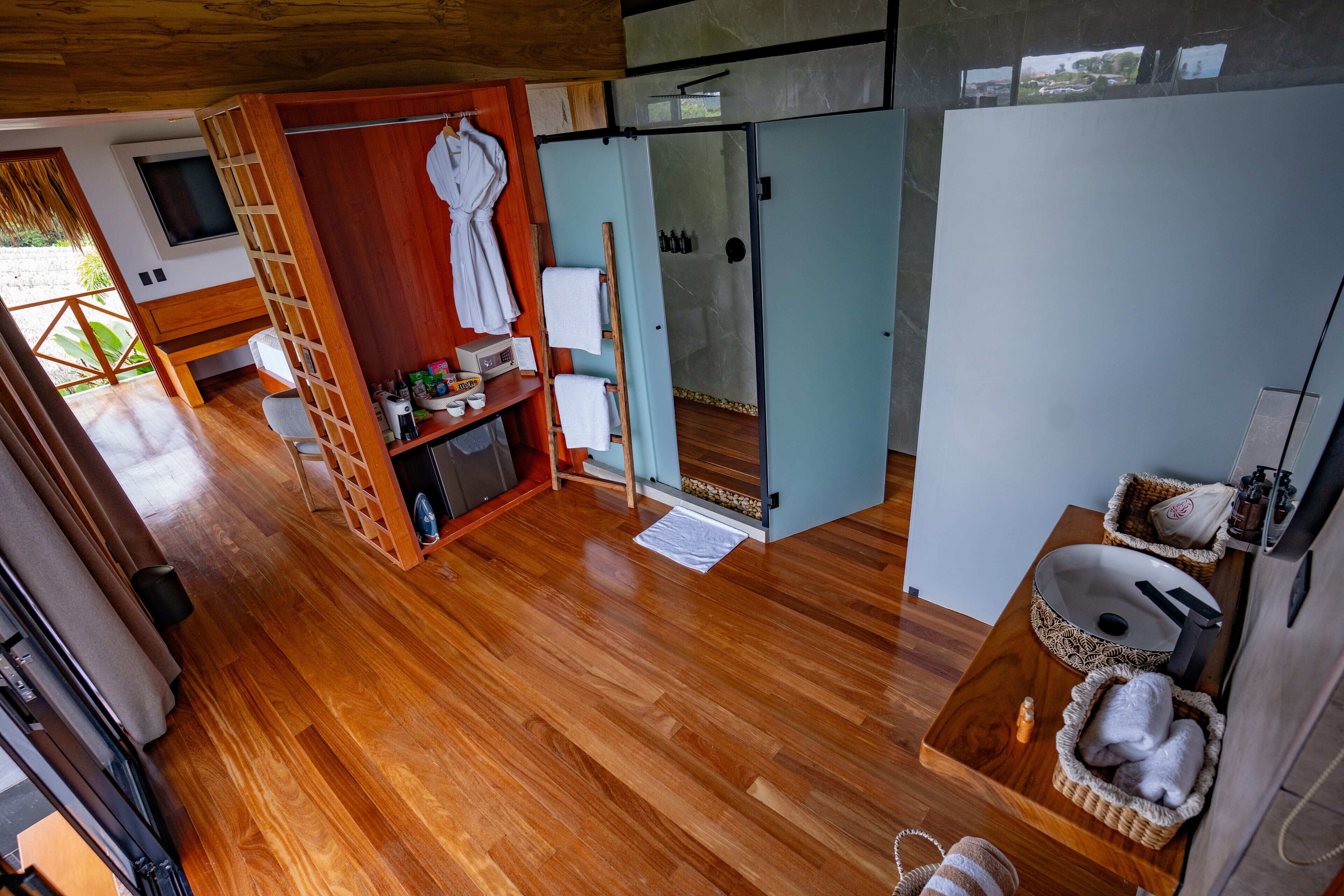 Habitación compacta con piso de madera, un pequeño rincón de descanso, baño con lavamanos y toallero, y vista al exterior.