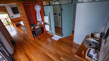 Habitación compacta con piso de madera, un pequeño rincón de descanso, baño con lavamanos y toallero, y vista al exterior.