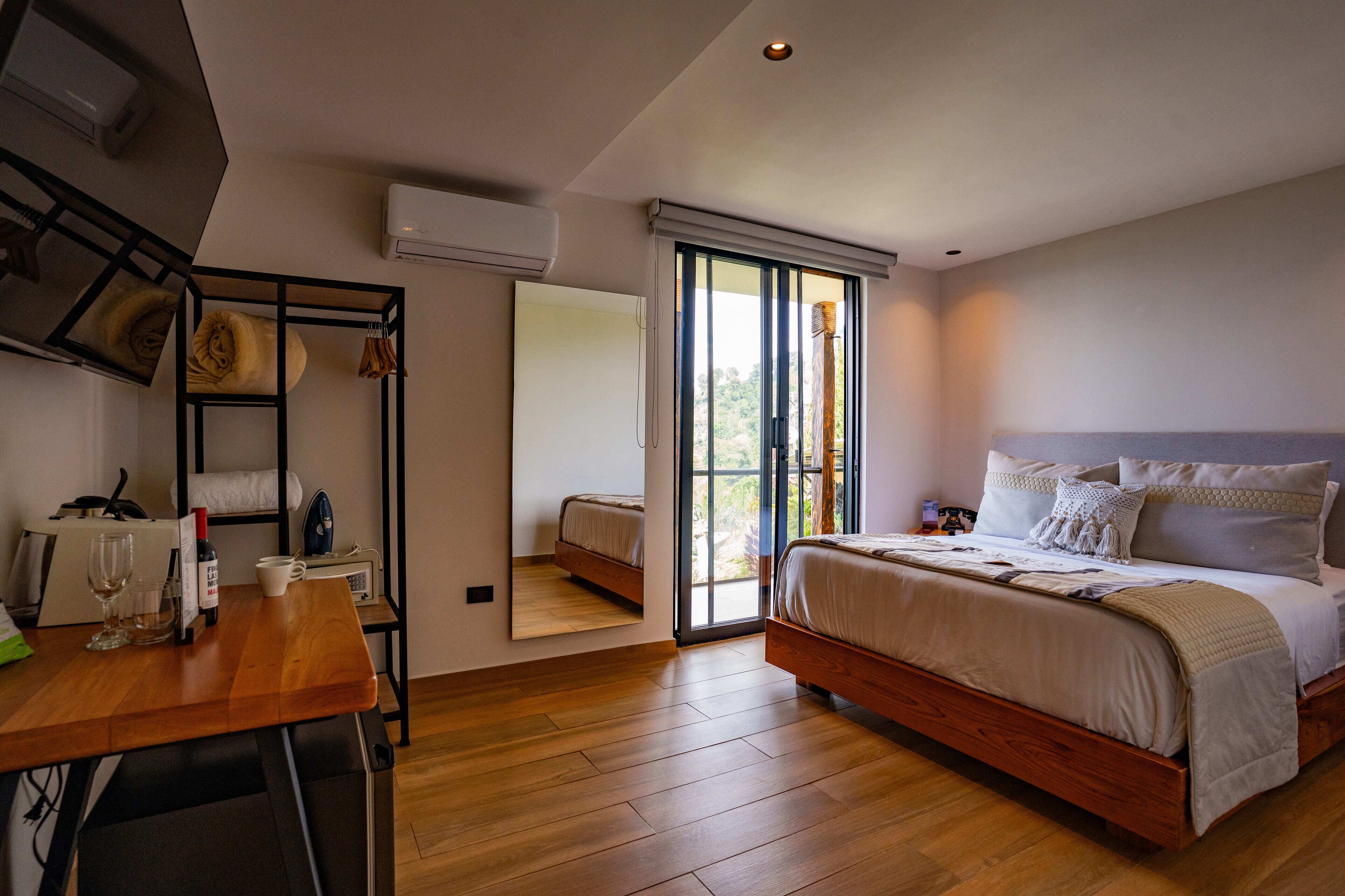 Une chambre avec un plancher en bois, un lit avec une literie blanche, un bureau avec une chaise, un miroir et une fenêtre avec des stores.
