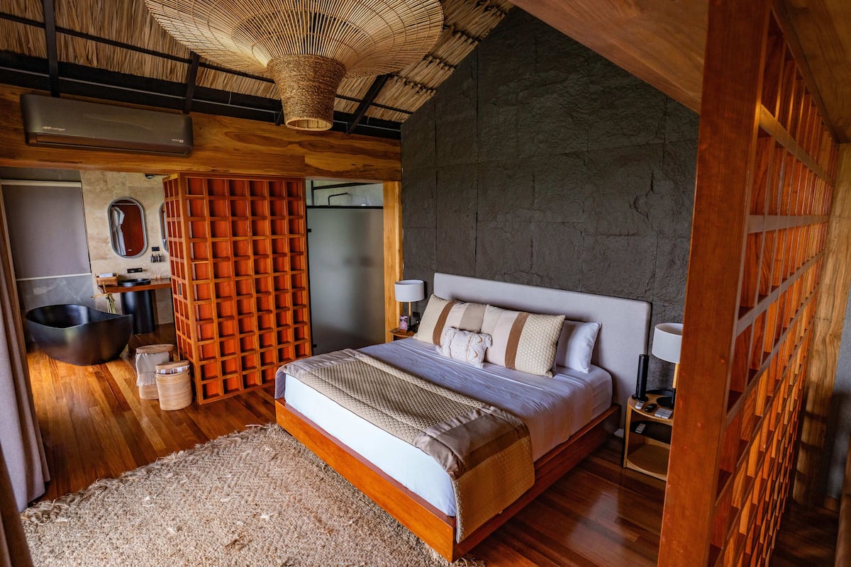 A bedroom with a large bed, a wooden floor, a stone wall, and a woven ceiling light fixture.