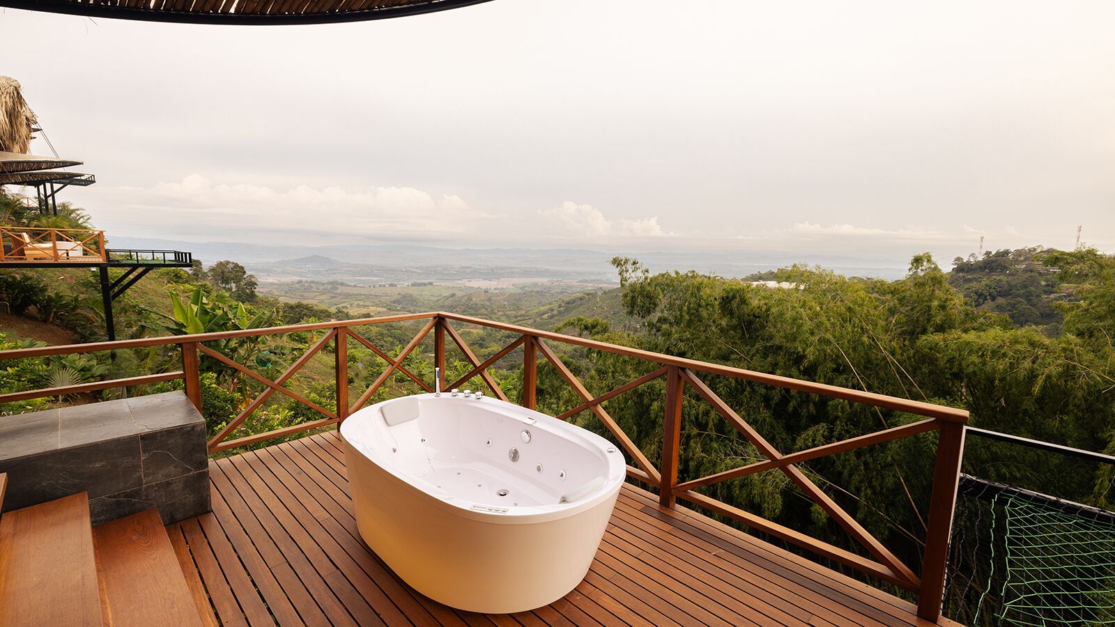 A wooden deck with a hot tub, a wooden table, and a railing overlooking a lush landscape.