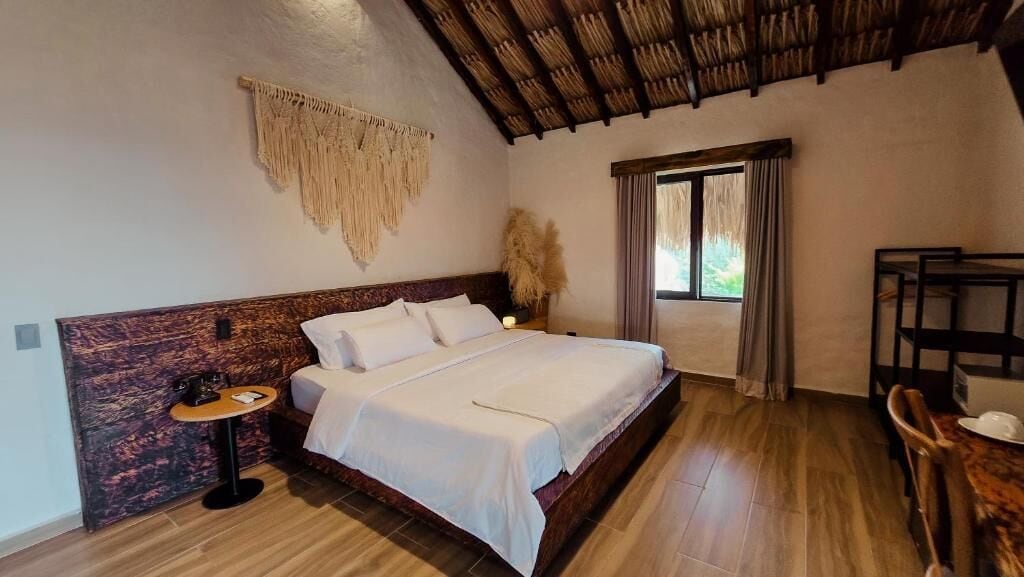 A bedroom with a bed, a round table, a chair, a window with curtains, and a macrame wall hanging.