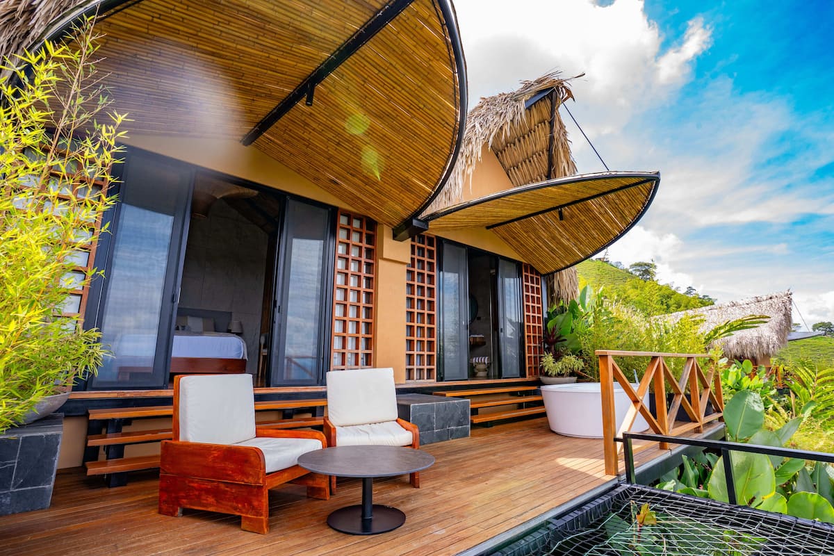 A wooden deck with outdoor seating, a thatched roof, and a view of lush greenery.