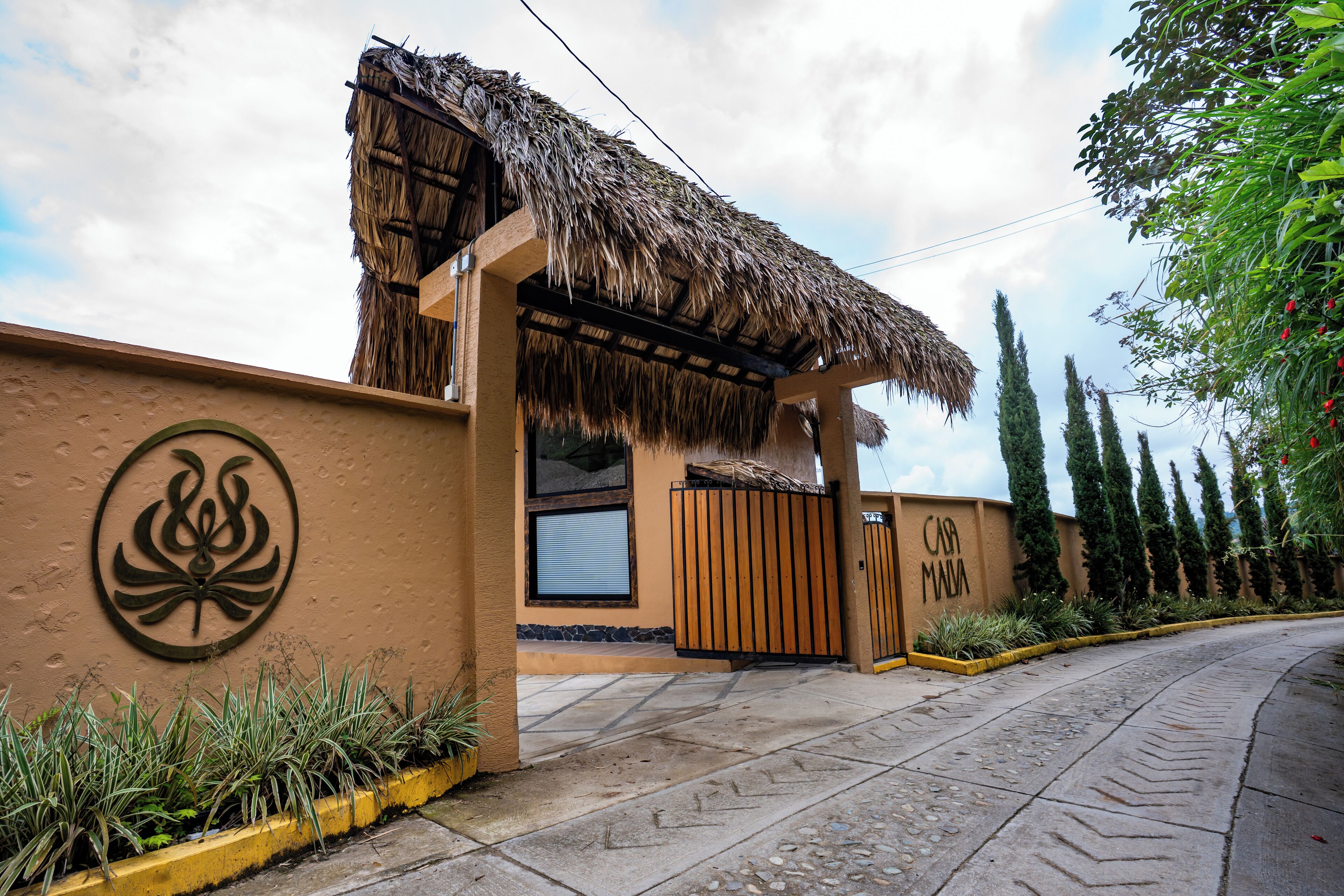 A building with a thatched roof entrance, a wall with a circular emblem, and a gated driveway.