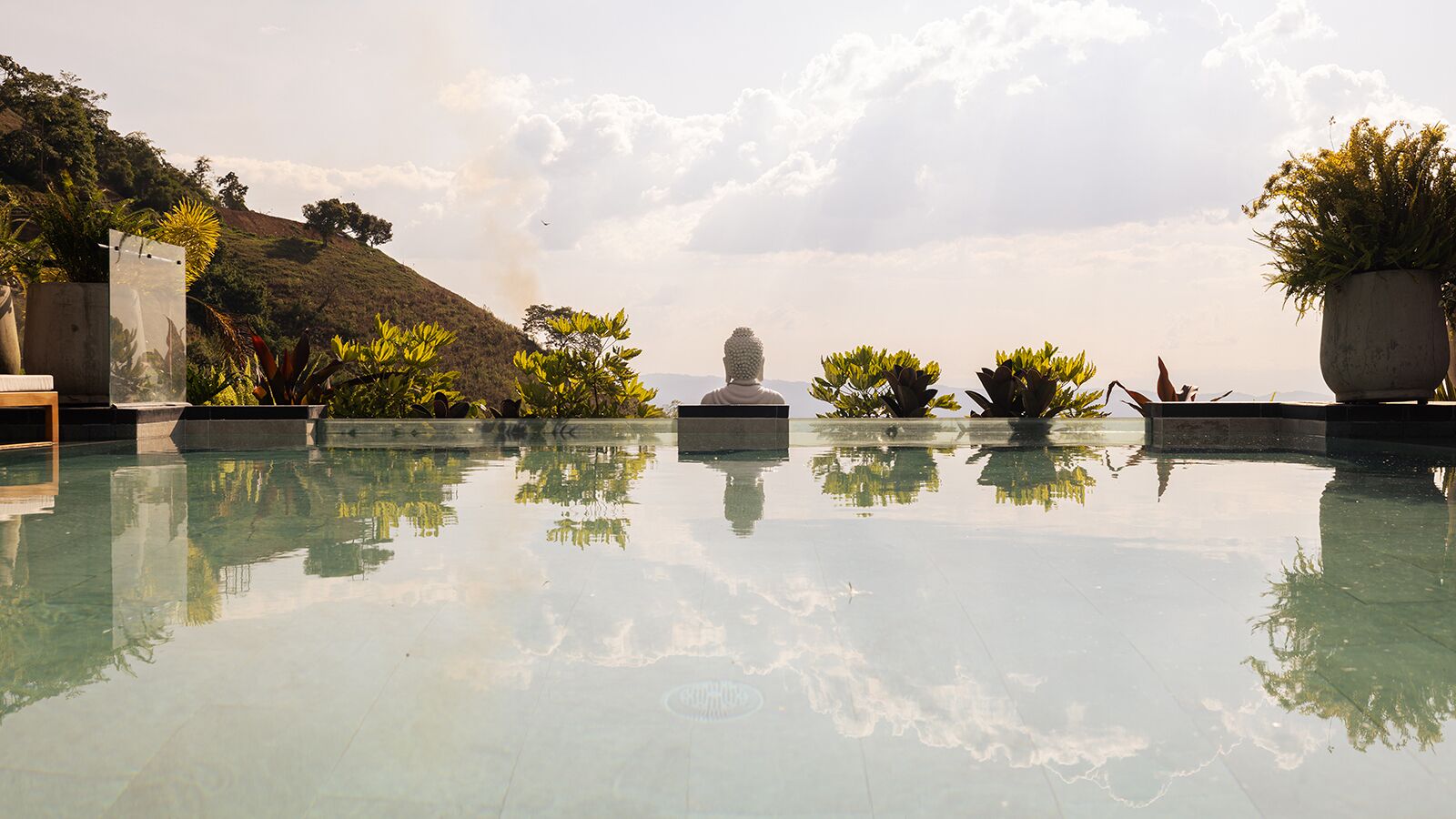 A serene infinity pool with clear water reflecting the surrounding landscape, including a statue and lush greenery.