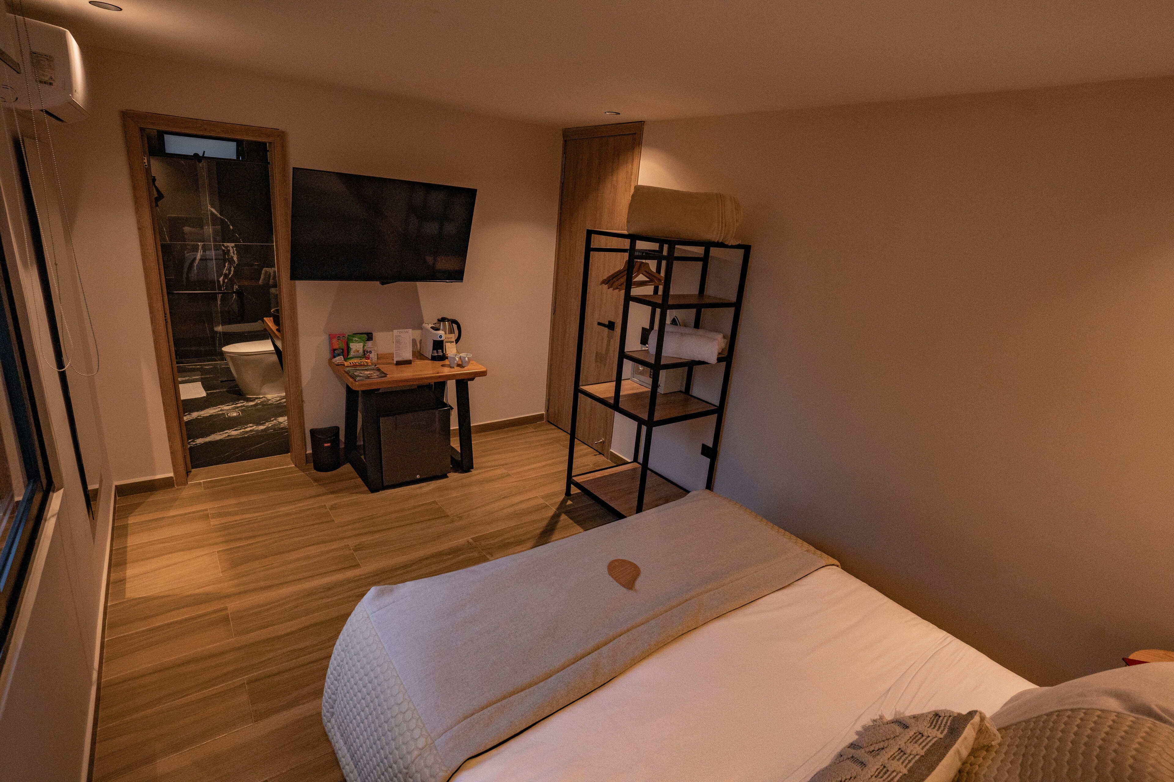 A hotel room with a bed, a TV mounted on the wall, a wooden desk with a chair, a small table, and a shelf unit.