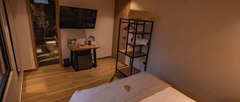 A hotel room with a bed, a TV mounted on the wall, a wooden desk with a chair, a small table, and a shelf unit.