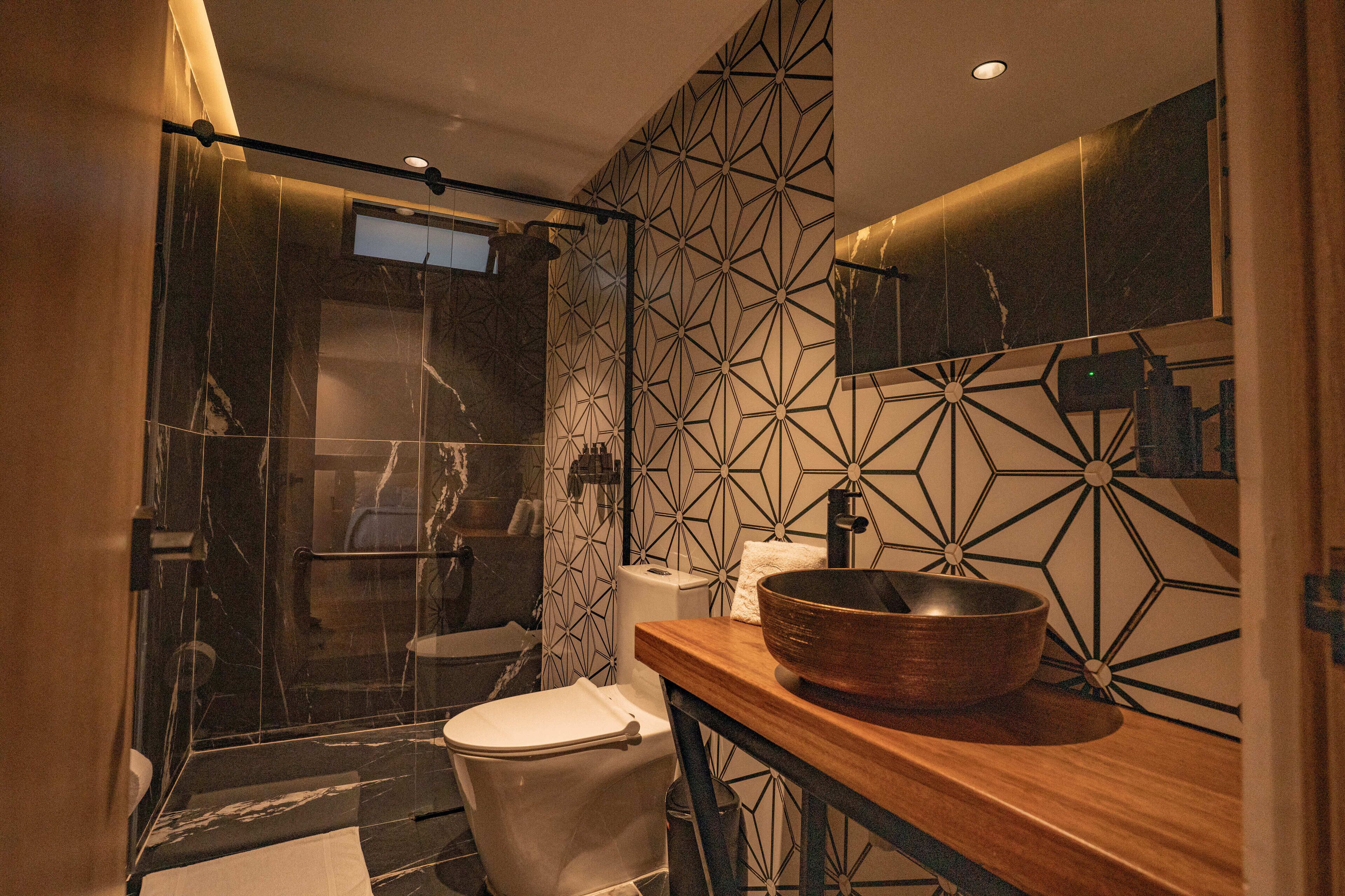 Un baño moderno con ducha envuelta en vidrio, inodoro blanco y tocador de madera con un lavabo grande de cobre.