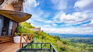 Una terraza de madera con una estructura de techo de paja, una maceta blanca y vistas a un valle exuberante.