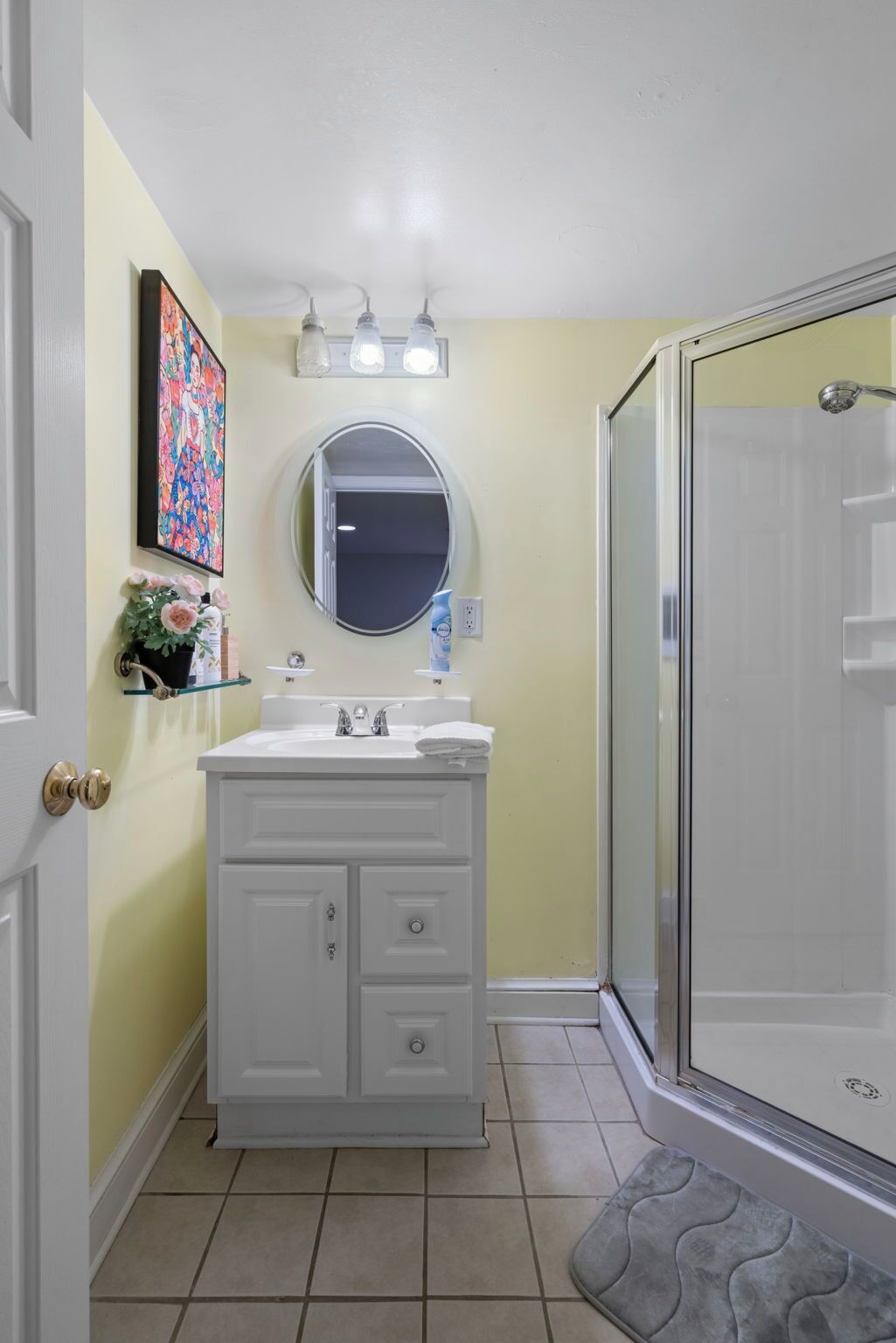 Een badkamer met een wit wastafelmeubel, een ronde spiegel, een douche met glazen deur en een betegelde vloer.