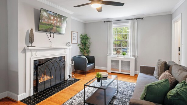 A living room with a fireplace, a ceiling fan, a television, a sofa, and a window with curtains.