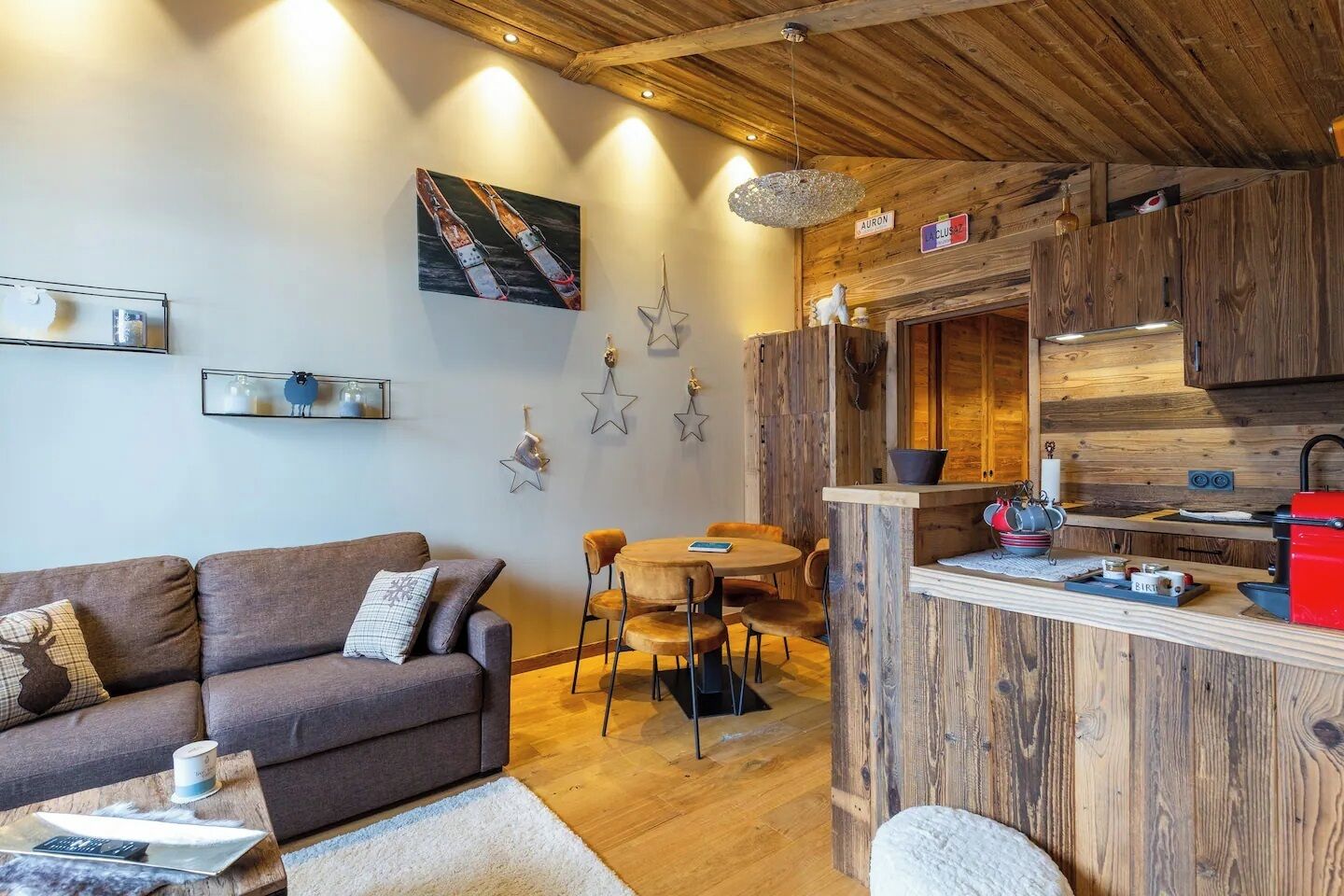 A cozy living area with a sofa, coffee table, and a dining table with chairs. A kitchen area with wooden cabinets and a red appliance is visible.
