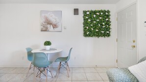 Un coin repas avec une table ronde, deux chaises de couleur turquoise, une petite plante en pot et une œuvre d’art florale encadrée accrochée au mur.