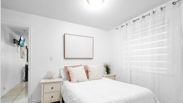 A bedroom with a white bed, a nightstand, a framed picture on the wall, and a window with sheer curtains. - Mountain View Italian Villa (Vancouver)