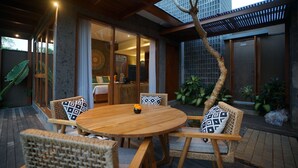 Une table à manger en bois, accompagnée de quatre chaises, un petit vase avec des fleurs et une vue sur un bâtiment moderne doté d’un toit en pente et de grandes fenêtres.