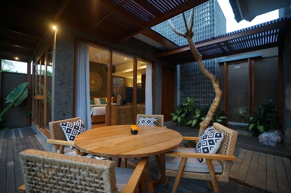 A wooden dining table with four chairs, a small vase with flowers, and a view of a modern building with a sloped roof and large windows. - The Shanti Graha Villas (Sukawati)