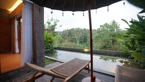 Une terrasse en bois avec deux fauteuils de jardin en osier, un grand parasol et une vue sur une étendue d’eau bordée de verdure.