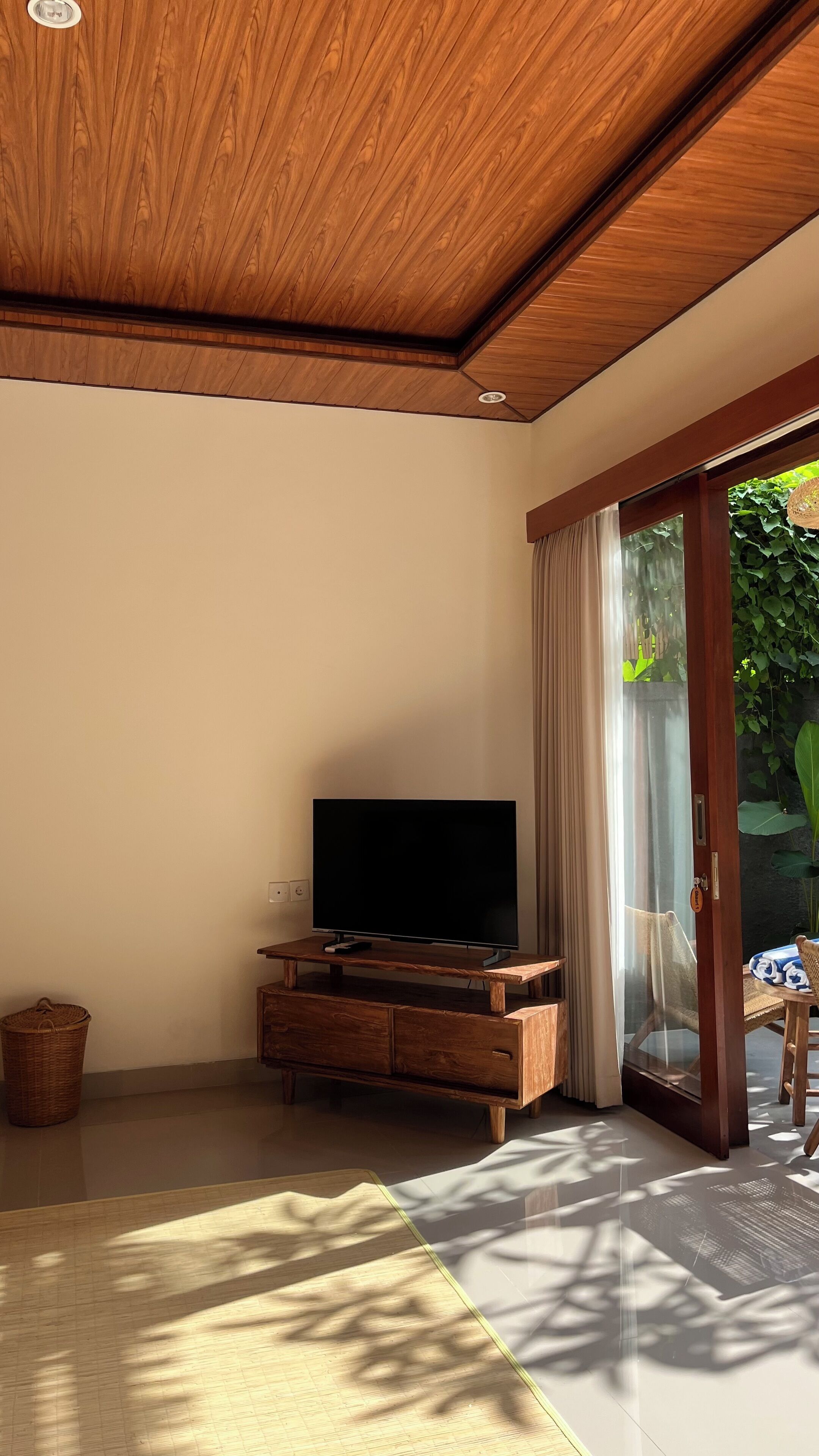 A room with a wooden ceiling, a flat-screen TV on a wooden cabinet, a sliding glass door to a garden, and a woven basket on the floor.