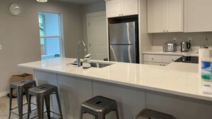 A modern kitchen with white cabinets, a stainless steel refrigerator, a large island with stools, and pendant lighting. - Kamal's Comfy Relaxation Zone (Eugene)