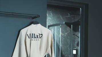 A white robe with "Villa37" text hanging on a wall, a pair of shoes on a small table, and a glass shower door.