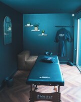 Un cuarto de terapia de masajes con piso de madera, una camilla, una silla de masaje, una ventana y un estante en la pared con plantas.