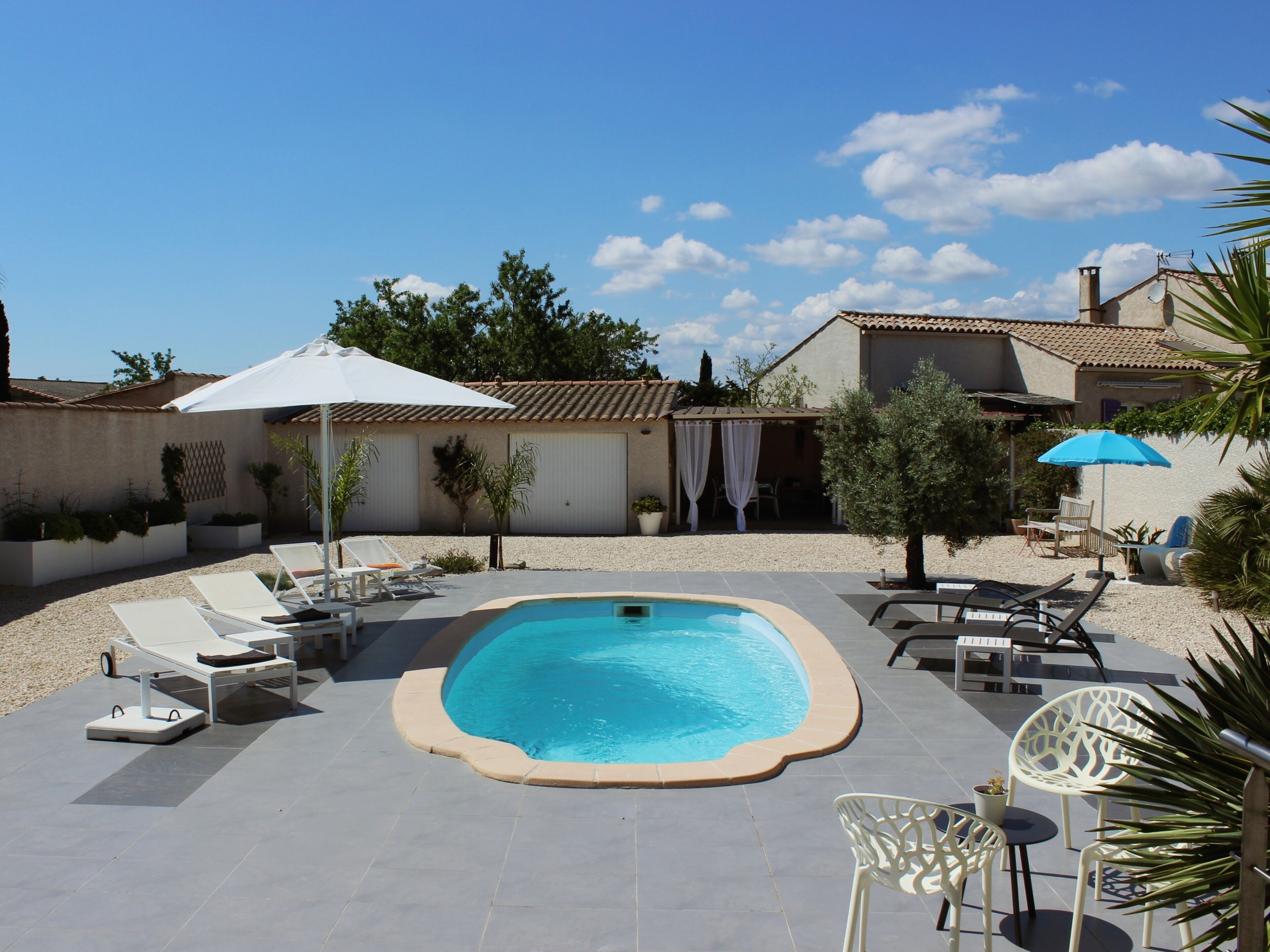 Ein Schwimmbecken mit Liegestühlen und Sonnenschirmen, umgeben von einer Terrasse mit Pflanzen und einem Gebäude im Hintergrund.