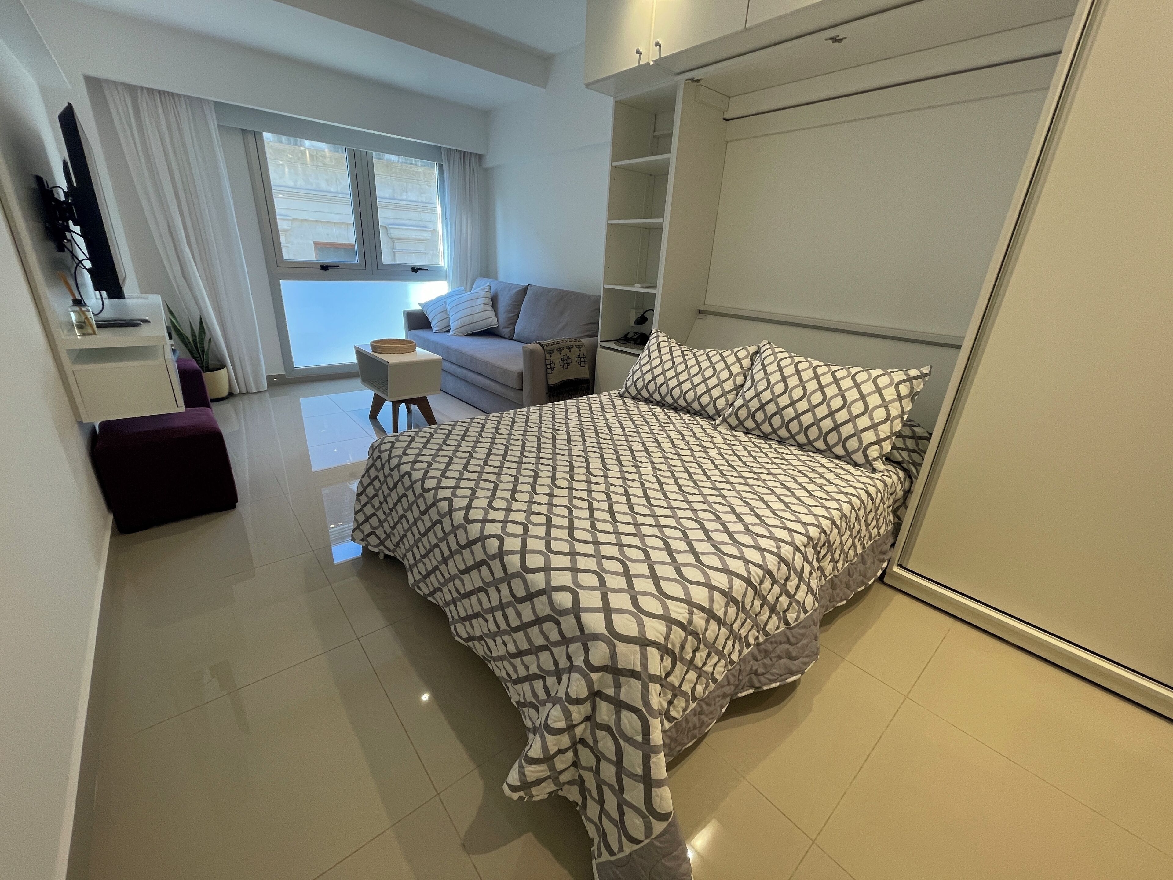 A modern bedroom with a large bed, a small desk, a sofa, and a window with curtains.