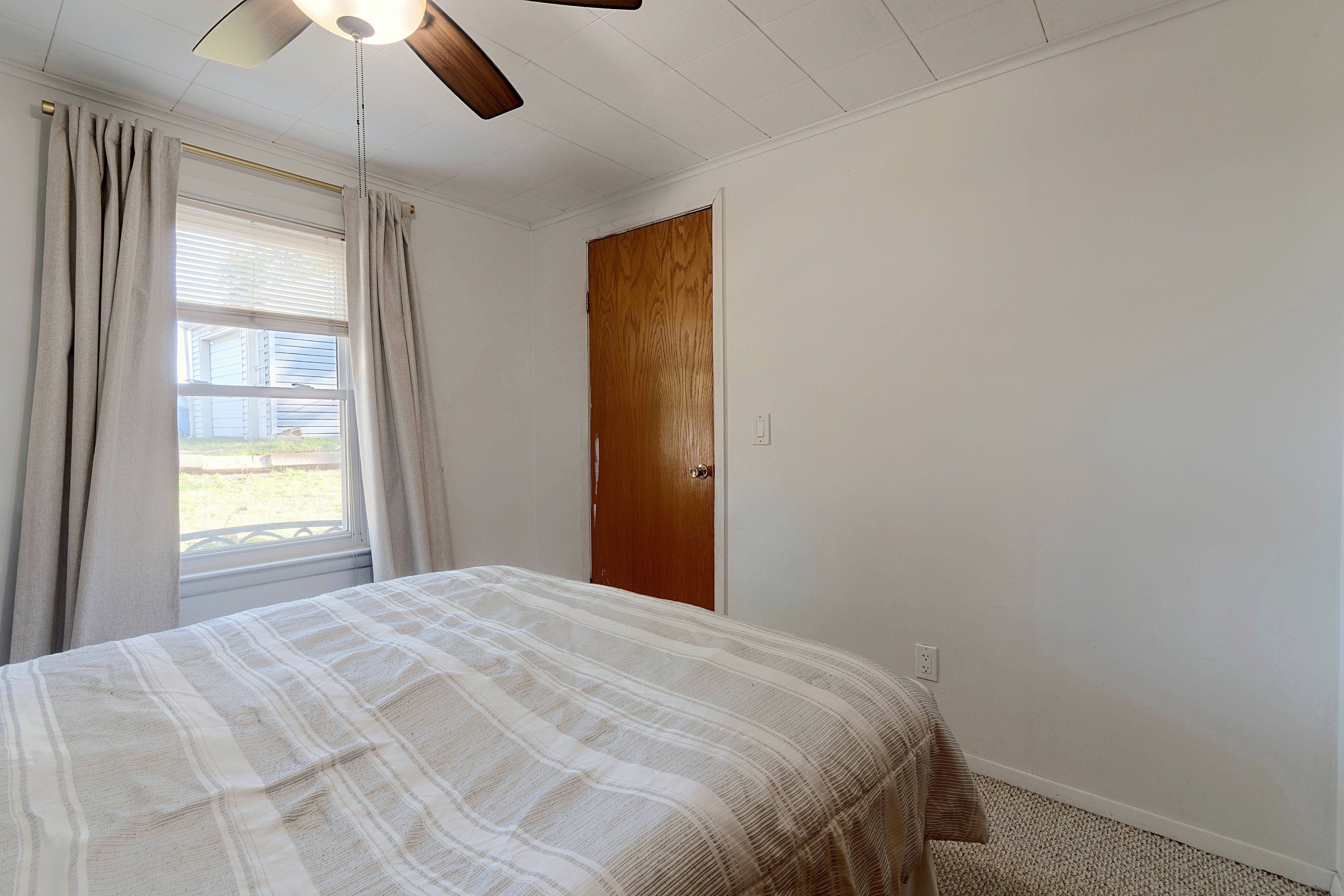 A bedroom with a bed, a ceiling fan, a window with curtains, and a wooden door.