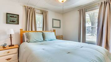 A bedroom with a large bed, a nightstand with a lamp, a framed picture on the wall, and a window with curtains.