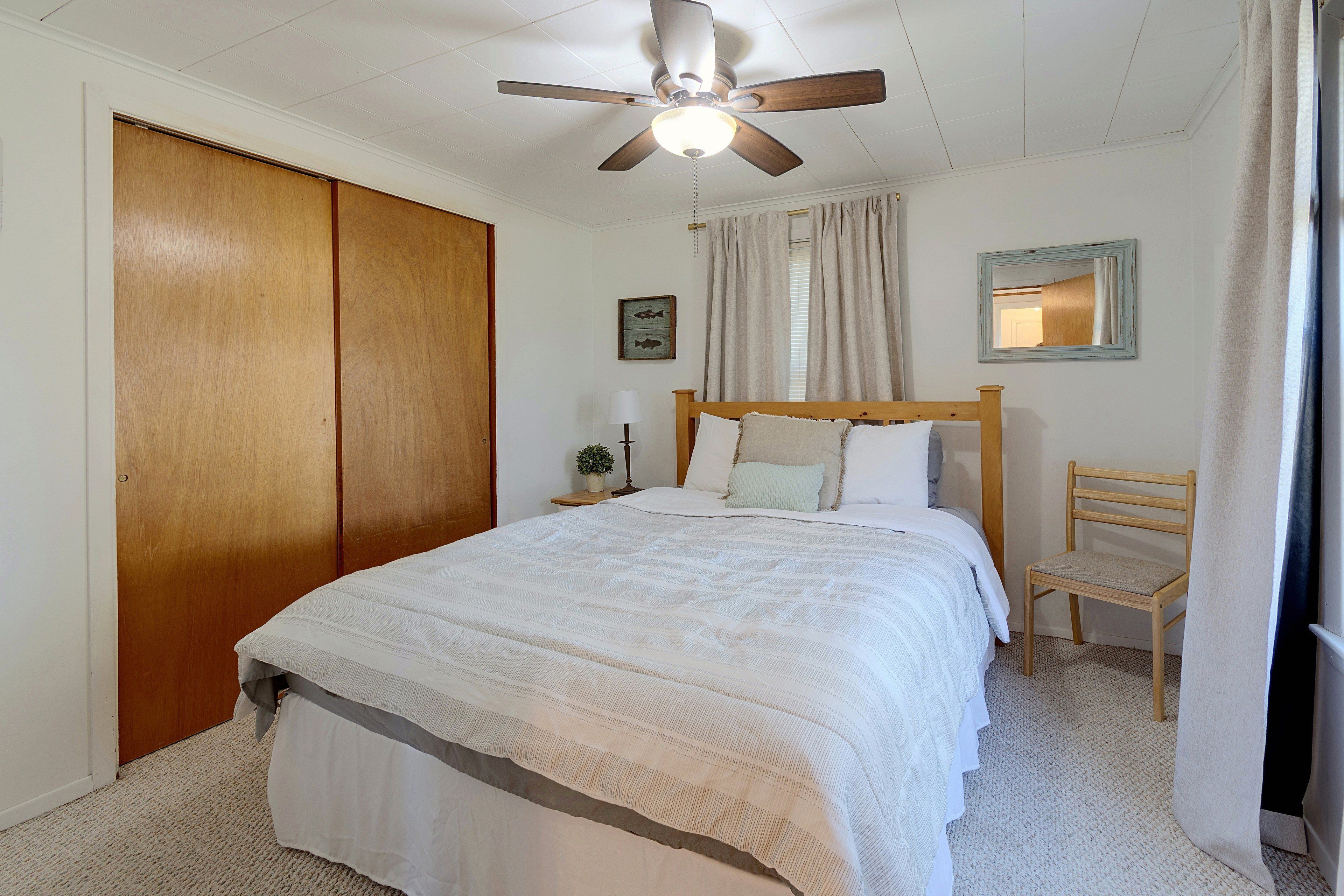 A bedroom with a bed, a wooden chair, a ceiling fan, a nightstand, a lamp, a framed picture, and a window with curtains.