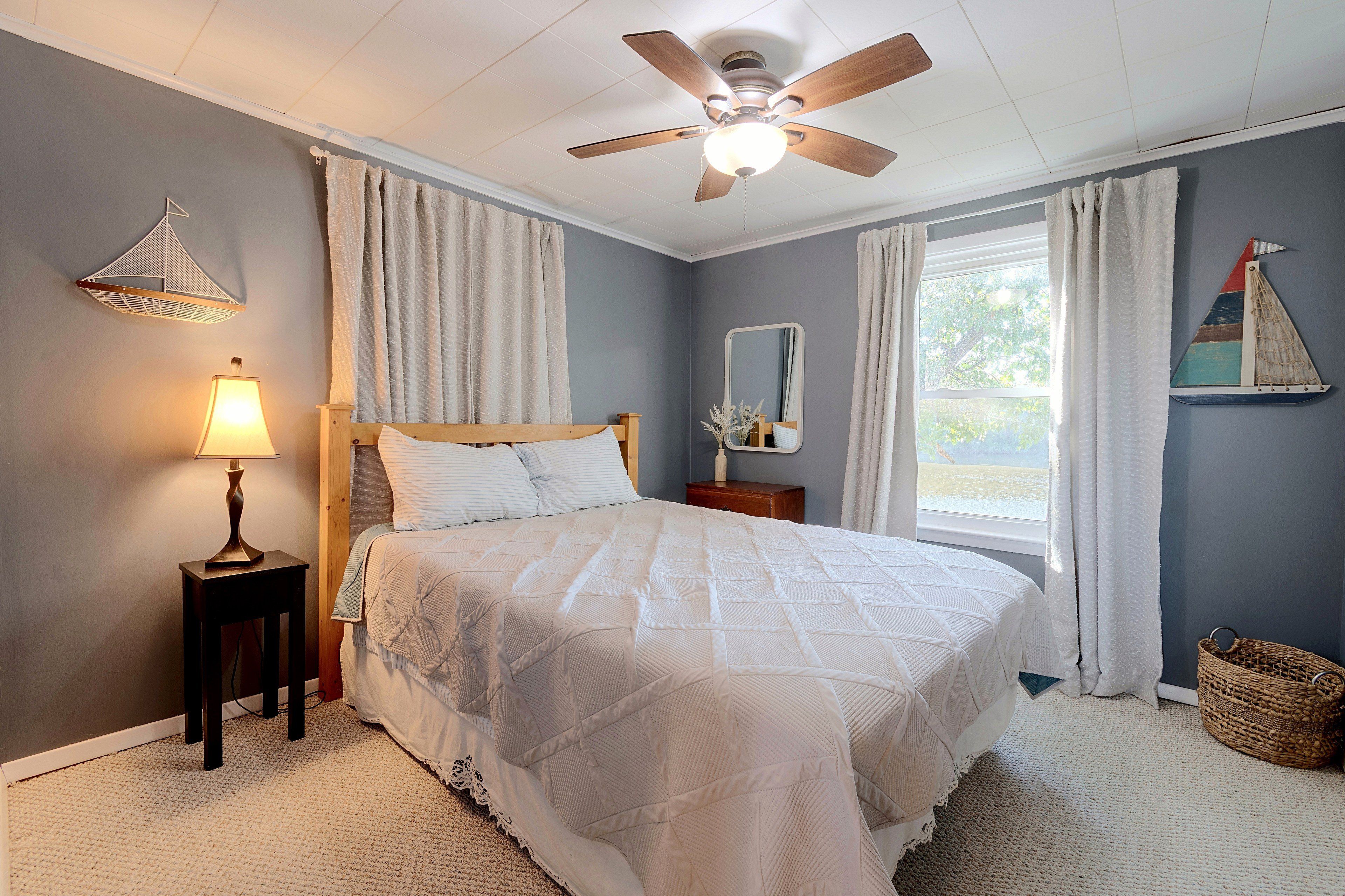 A bedroom with a bed, a ceiling fan, a nightstand, a lamp, a window with curtains, and a wall-mounted sailboat decoration.