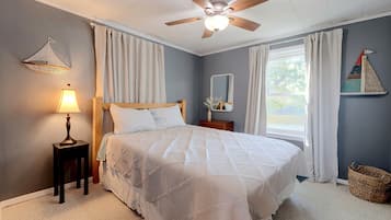 A bedroom with a bed, a ceiling fan, a nightstand, a lamp, a window with curtains, and a wall-mounted sailboat decoration.