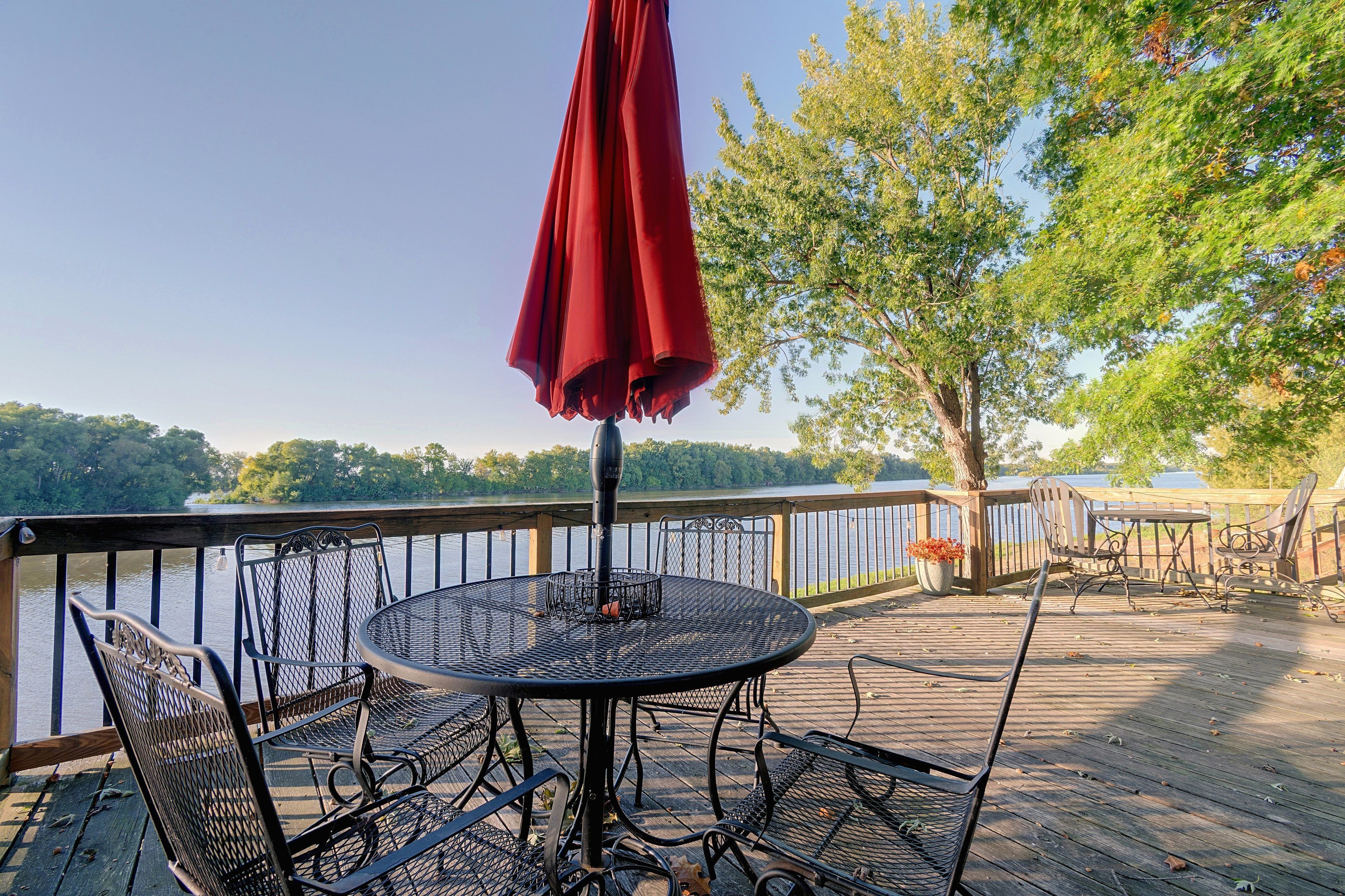 A deck with a round table and chairs, a red umbrella, and a view of a lake and trees.