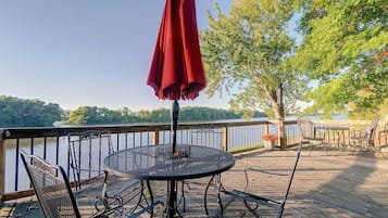 A deck with a round table and chairs, a red umbrella, and a view of a lake and trees.