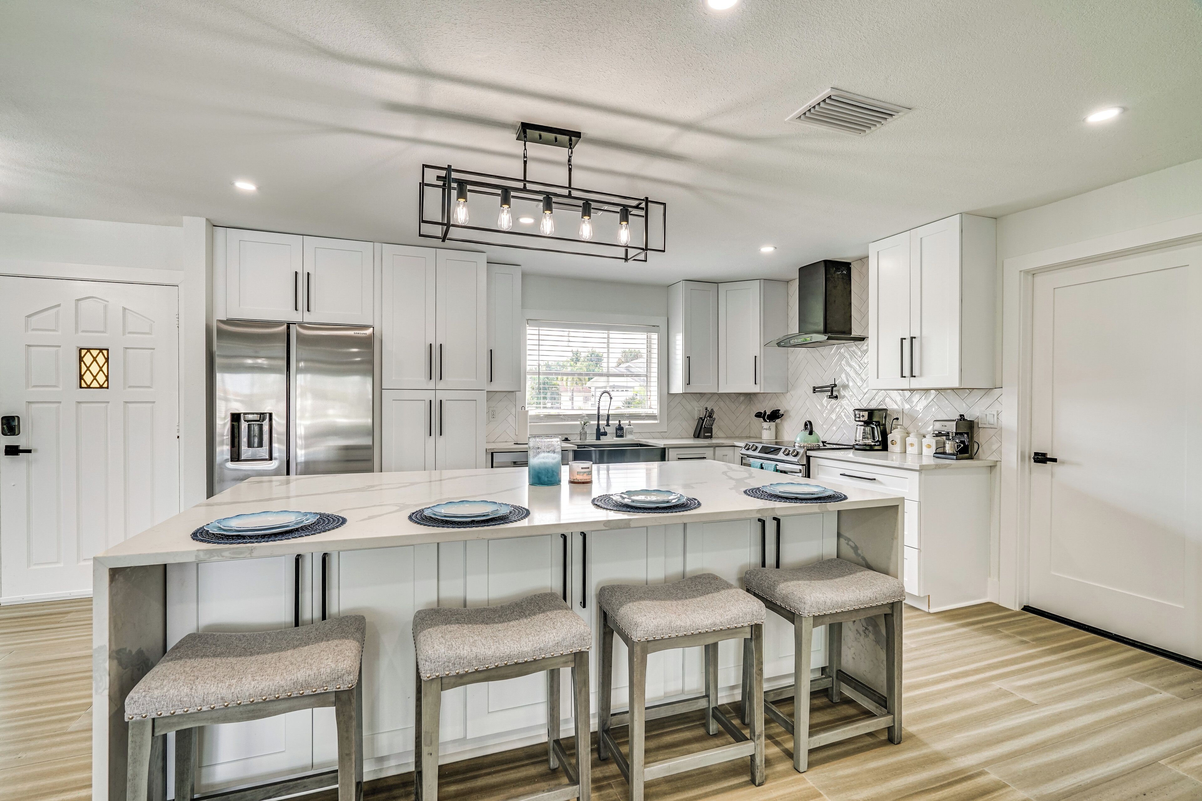 A modern kitchen with white cabinets, a stainless steel refrigerator, a central island with bar stools, and a pendant light.