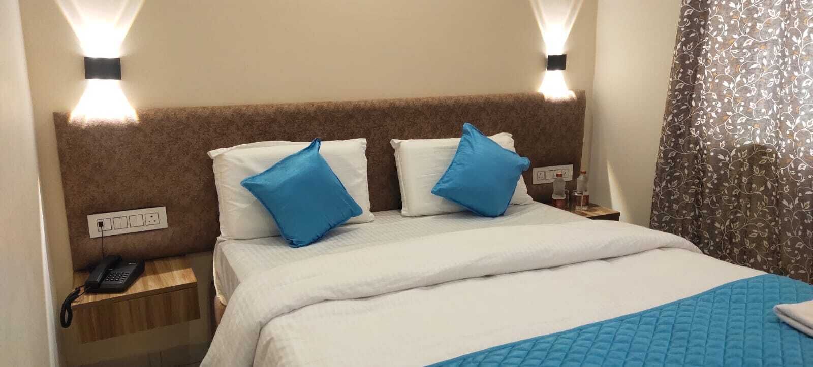 A hotel room with a bed, two pillows, a bedside table with a phone, and a wall-mounted light fixture.