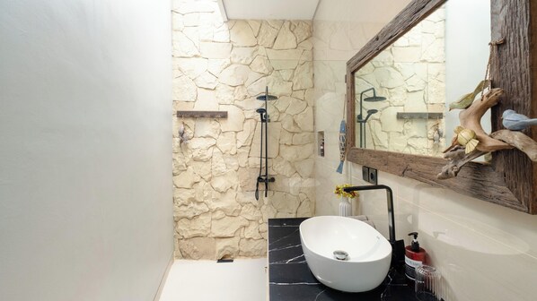 A bathroom with a stone wall, a white sink, a wooden shelf, and a mirror with a rustic frame.
