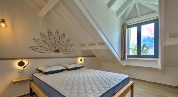 Een slaapkamer met een schuine plafond, een bed met een witte matras en een blauwe spreidek, twee nachtlampjes en een decoratief kunstwerk van bladeren aan de muur.