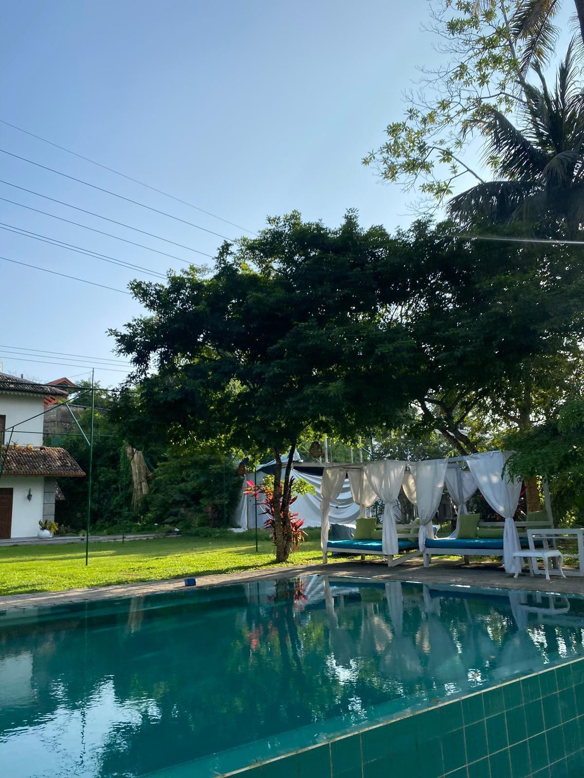 Una piscina amb una estructura blanca que fa de tanca i zona de seients, envoltada de vegetació abundant i una casa al fons.