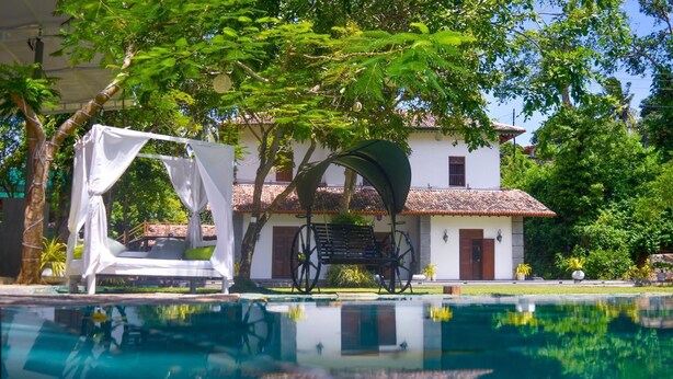 Une piscine surmontée d’un auvent blanc et bordée d’un banc, le tout entouré d’une végétation luxuriante et d’un bâtiment blanc doté d’un toit en tuiles.