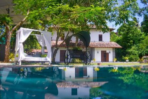 Una piscina con un toldo blanco y una banca, rodeada de exuberante vegetación y un edificio blanco con techo de tejas.