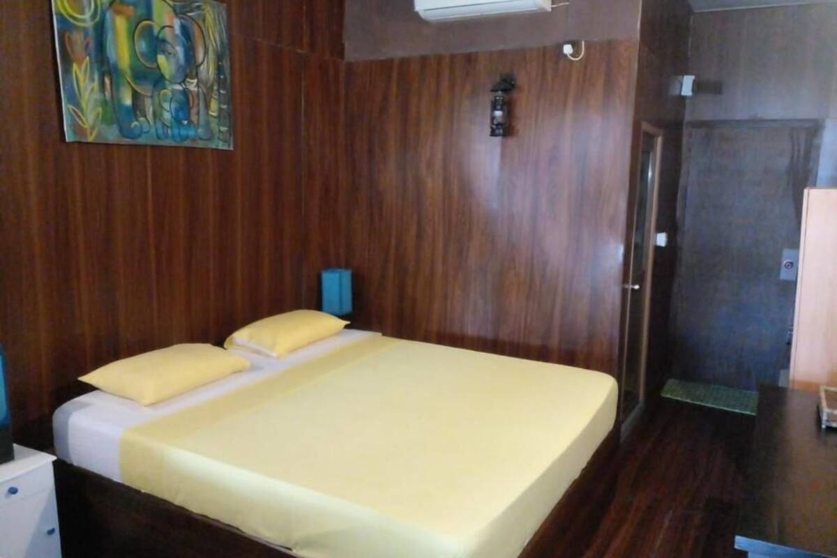 A room with a wooden wall, a bed with yellow bedding, a small bedside table, and a painting on the wall.
