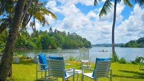 Une vue sur le lac, avec des fauteuils à bascule blancs et une table, le tout entouré d’une végétation luxuriante et de palmiers.