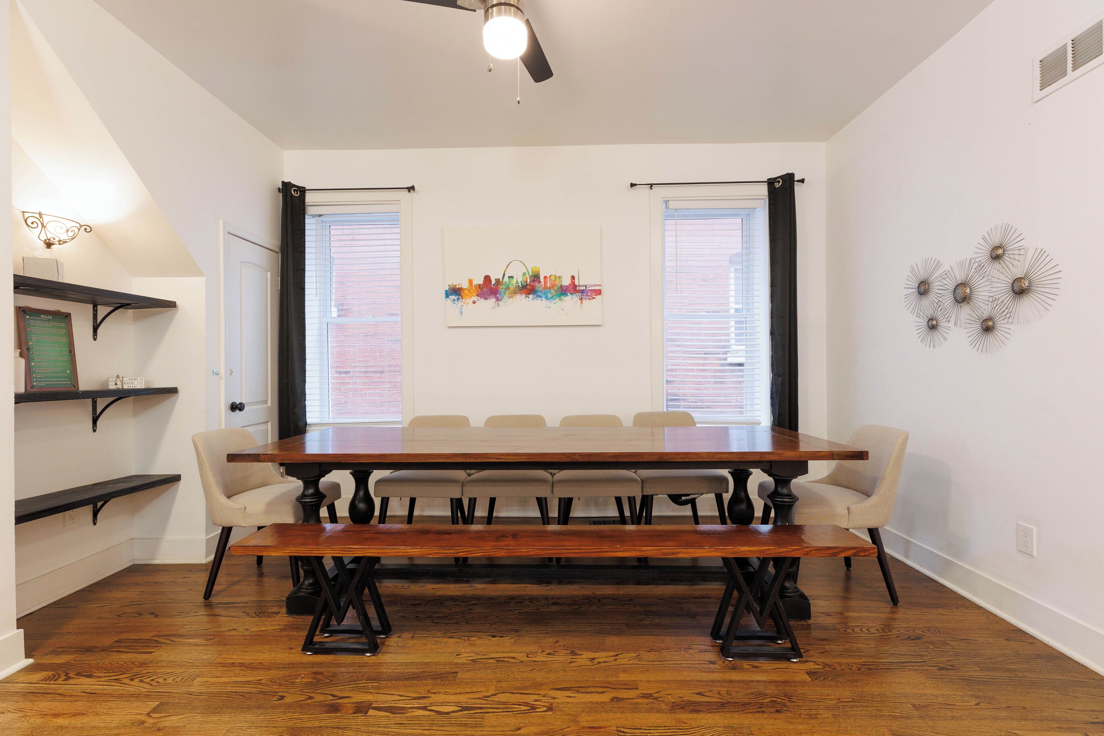 A dining area with a wooden table and benches, two windows with blinds, a ceiling fan, and wall-mounted decor.