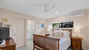A bedroom with a wooden bed, two bedside lamps, a ceiling fan, a TV, and a window with a view of greenery.