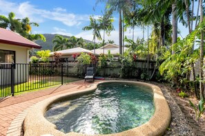 Un patio trasero con piscina, patio de ladrillo y área cercada con palmeras y otra vegetación.